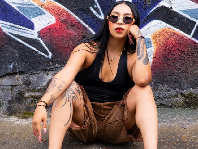ChloeMoretti, squatting against a graffiti-covered wall. She wears a black sleeveless top, brown frayed shorts, and sunglasses. Her long black hair flows over her shoulders, and she has tattoos on her right arm and left thigh. Her right hand touches her face, and her left arm rests on her knee. Her red lipstick contrasts with her tan skin. The graffiti features bold colors: red, blue, and white. The concrete ground shows wear. She's accessorized with a bracelet and necklace. Her nails are painted red.