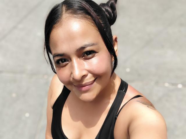 AfroditaLaDiosa, a young Asian woman with medium skin tone and black hair in a small bun, looks up at the camera with a slight smile. She has a subtle tattoo on her right shoulder. She wears a black tank top with a low neckline. The image is a close-up with natural light highlighting her smooth skin and slightly flushed cheeks. Her eyebrows are dark and well-defined. The background is a gray, textured pavement. The composition is centered on her face and upper torso.