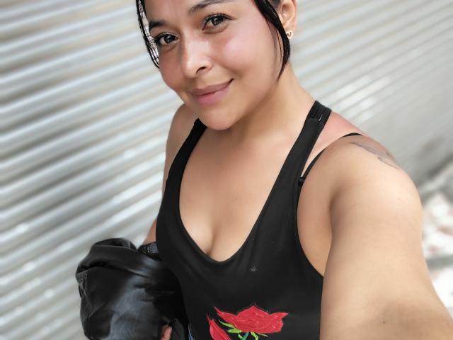 AfroditaLaDiosa, a young woman with light brown skin and dark hair in a simple updo, smiles at the camera. She wears a black tank top with a red rose graphic and holds a black leather jacket. A small tattoo is visible on her right shoulder. Her brown eyes and subtle makeup highlight her natural beauty. The image has a casual, street-style vibe. The focus is on her upper body and face.