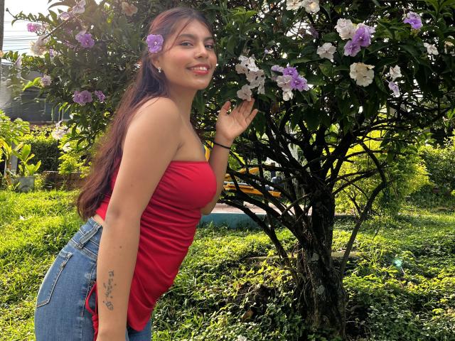 AlyssaGreen stands in a green garden, leaning slightly forward with a hand raised. She has long, dark brown hair, a small tattoo on her right forearm, and wears a red strapless top and blue jeans. She's smiling at a tree with purple and white flowers. Sunlight filters through the leaves, highlighting her skin and the flowers. The image has a casual, natural feel.