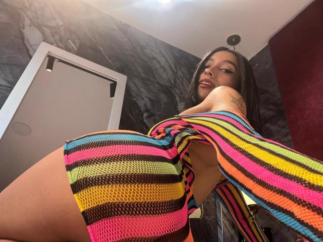 SalomeMille with a big ass, wearing a colorful striped dress, looking down at the camera with a tattoo on her right arm, in a modern room with dark walls.