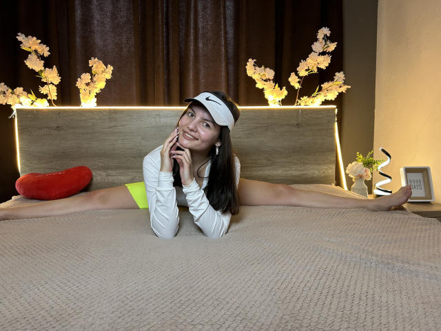 JasmineAsha is doing a full split on a bed, wearing a white top, neon yellow skirt, and white headband, with arms raised, smiling, and showing off her flexible ass.