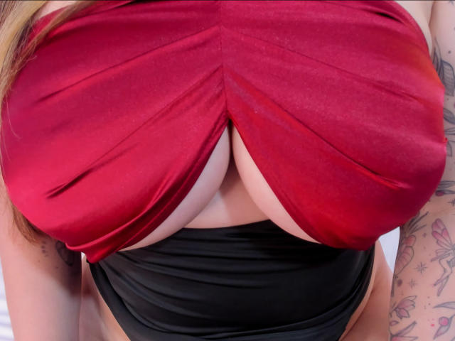 Close-up of ChristineBray's chest, showing her large, round breasts partially covered by a red, crisscrossed fabric that accentuates her cleavage. Below, a black, strapless garment is visible. Her light skin contrasts with the red fabric. Her right arm, with a tattoo of flowers and stars, is slightly visible on the right. The image focuses on the texture of the fabric and the curvature of her breasts. The red fabric is smooth, and the black garment is sleek. The tattoos add a colorful, detailed element to the composition.