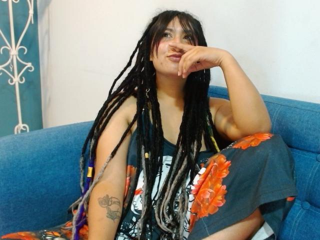BeckyHuston sits on a blue sofa, her long black dreadlocks with colorful extensions flowing down her shoulders. She wears a dark gray t-shirt with a graphic design and a floral-patterned cover-up. Her right arm rests on the sofa, her fingers touching her chin. She has a tattoo on her left forearm. The room has a white wall and a teal curtain with a white decorative pattern. Her expression is relaxed, and she looks slightly to the right. The image has a casual, everyday vibe.