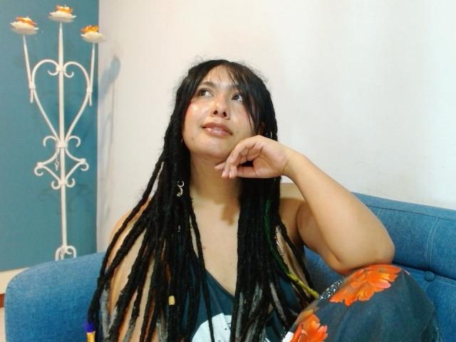 BeckyHuston, a young woman with dark, thick dreadlocks, sits on a blue sofa. She has a light brown skin tone and looks upwards with a slight smile. She wears a black sleeveless top with a white graphic and has orange floral patterns on her right sleeve. Her left hand rests on her chin. The background shows a white wall and a decorative, white metal stand with three small, orange-topped flower-like ornaments. The image has a casual, homey feel.