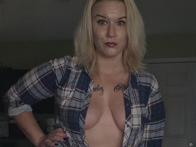 RhondaRump stands in a dimly lit room, facing the camera. She has blonde hair, wearing an unbuttoned plaid shirt, exposing her large, natural breasts with visible nipples. She has tattoos on her chest reading "Lily" on the right side and another tattoo below her collarbone. Her right hand rests on her hip, and she has dark red lipstick. The image is a photograph with a casual, slightly grainy texture. RhondaRump's confident, direct gaze is focused on the camera.