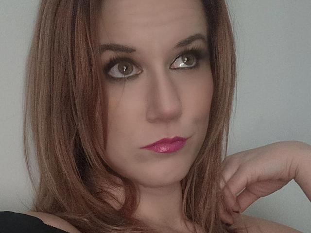 RhondaRump's face with blue eyes, blonde hair, and a subtle smile. She's wearing black eyeliner and has a small nose with slightly parted pink lips. Her boobs are partially visible.