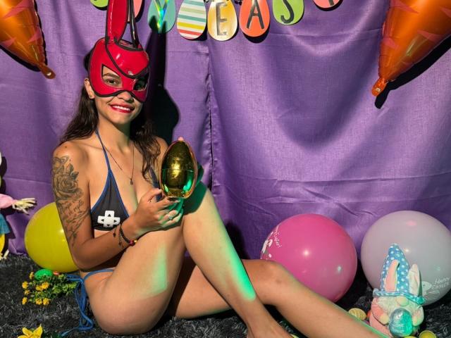 CanelitaRousse sits on a black furry surface, wearing a red mask with rabbit ears and a blue string bikini. She holds a shiny gold egg in her right hand. Her left arm has a detailed tattoo. Colorful balloons and a stuffed bunny with a blue polka-dot bow are in the background. Her long dark hair is loose, and she has bright red lipstick. The purple backdrop has "HAPPY EASTER" letters. The image uses green and purple light to highlight her legs. She smiles at the camera.