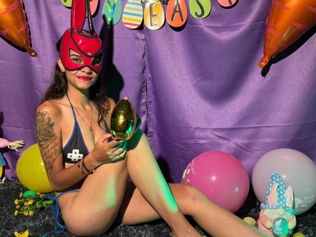 CanelitaRousse sits on a black carpet, wearing a red mask with bunny ears and a blue string bikini. She holds a shiny gold egg, with tattoos on her right arm. Her long brown hair is loose, and she has a slight smile. Background includes purple fabric, colorful balloons (yellow, pink, white), and a stuffed bunny. Bright green and yellow confetti is scattered on the carpet. The image has vibrant colors and a playful, festive atmosphere.