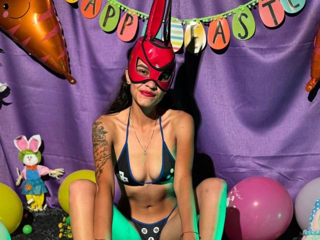 CanelitaRousse with red mask, dark blue bikini, showing her tits, tattoo on left arm, sitting on floor with balloons and Easter decorations, smiling, long brown hair.