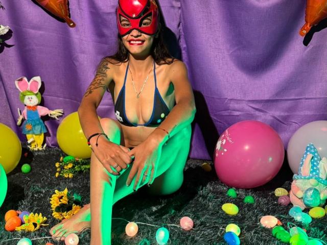 CanelitaRousse, wearing a red mask, dark blue bikini with yellow dots, sitting on a green carpet, surrounded by Easter eggs, balloons, and plush toys, smiling at the camera.