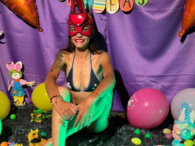 CanelitaRousse sits on a black fuzzy carpet, wearing a red mask with rabbit ears, a dark blue string bikini, and a silver necklace. She has a tattoo on her right arm and is smiling with red lipstick. Her legs are crossed, and she has green light on her left leg. Surrounding her are colorful balloons, a pink balloon with a white star, a small doll in a blue dress with a pink bow, and scattered Easter eggs. The purple backdrop has colorful letters. The image has a playful, festive vibe.