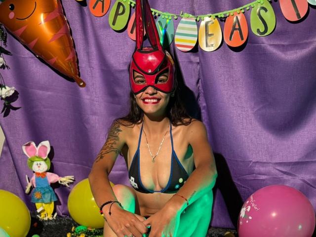 CanelitaRousse, a woman with medium brown skin and dark curly hair, sits on the ground in a brightly lit scene. She wears a red superhero mask, a dark blue bikini with white polka dots, and has a tattoo on her right arm. She's smiling with red lipstick. The background is a purple curtain with colorful "Happy Easter" letters and decorations including a green and pink balloon, a stuffed bunny, and a shiny orange Easter egg. Green and yellow lights illuminate her.