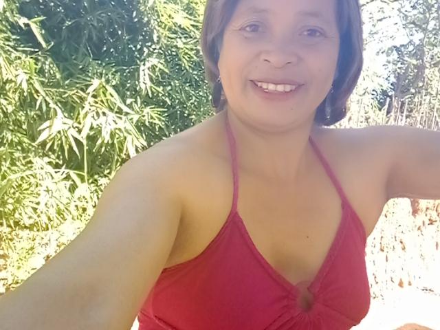 Rahel69 with short brown hair, smiling, wearing a red halter top that shows her small tits, sitting outdoors in sunlight. She has fair skin and is holding the camera.