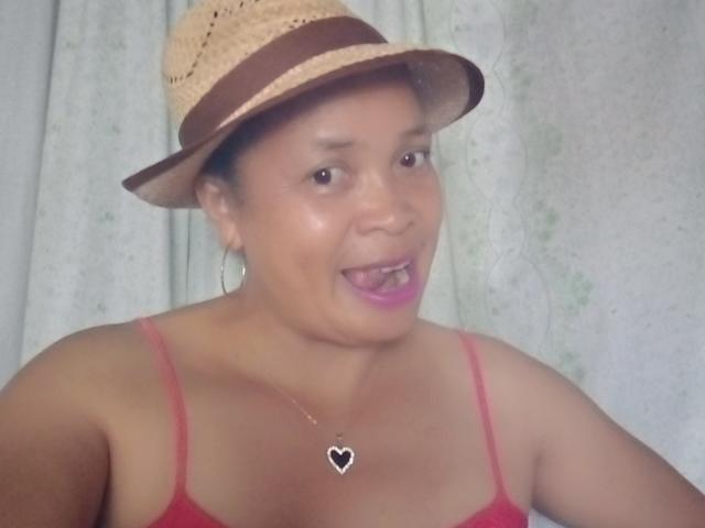 Rahel69 with tan skin, brown eyes, and pink lipstick, wearing a straw hat, red tank top, hoop earrings, and a heart necklace, mouth open in a surprised expression.