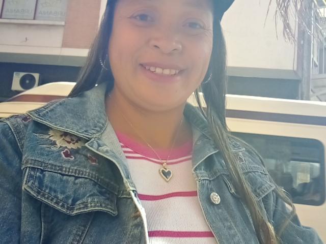 Rahel69, Latina with long brown hair, wearing a black cap, denim jacket with heart patch, white and pink striped shirt, heart necklace, smiling, showing small white teeth.