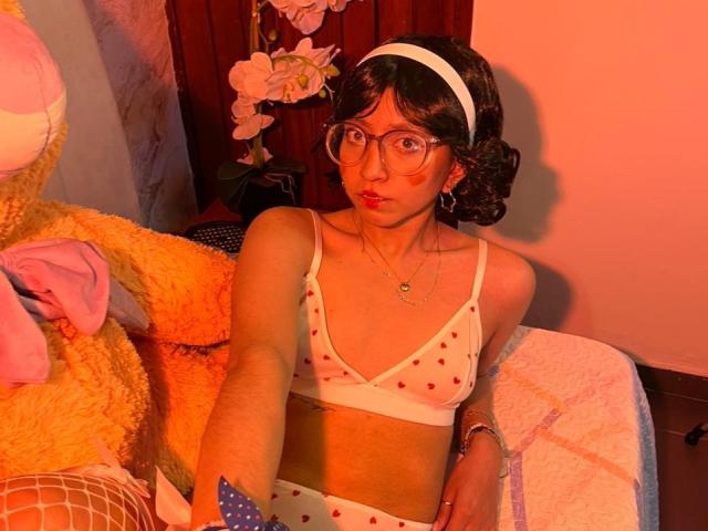 VioletXBloom with glasses, red lipstick, white headband, polka-dot bra and panties, holding a blue flag, sitting next to a giant yellow stuffed toy, in warm orange light.