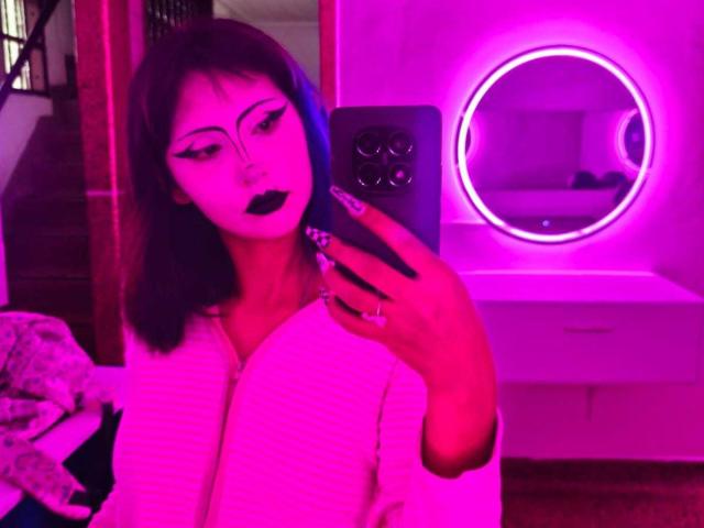 A selfie of KleoLennox with pink neon lighting. KleoLennox has dark blue and black hair, wearing a white ribbed sweater, and has bold black eyeliner, dark lipstick, and long manicured nails with black and white stripes. KleoLennox holds a black smartphone with a circular camera cutout in the back. A round mirror with a pink neon ring is in the background on a white dresser. The room has a pink neon glow.