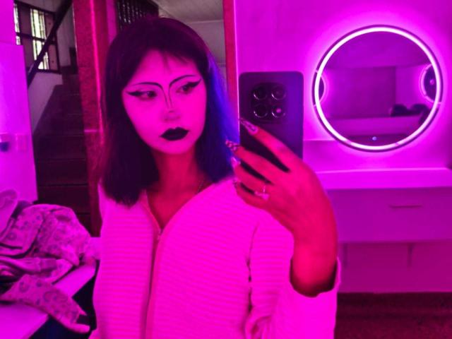 KleoLennox with dark blue and purple hair, white ribbed robe, black lipstick, and eyeliner, holding phone, taking mirror selfie, illuminated by pink and purple neon light.
