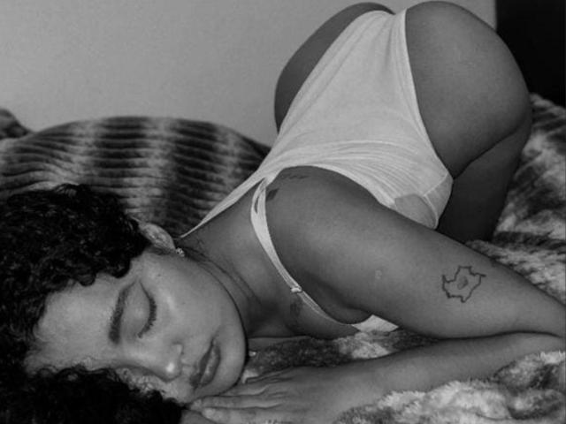 Black and white photo of MaddiAndAndrew with curly hair, lying on a fuzzy blanket. She's on all fours, wearing a white thong and tank top, with her eyes closed and a relaxed expression. Her tattoo, a simple line drawing of a star, is on her upper arm. Her skin is dark, and the texture of the blanket is rough. The composition highlights her curvy, round buttocks and the softness of her face. The image uses high contrast, emphasizing her relaxed posture and the texture of the blanket.