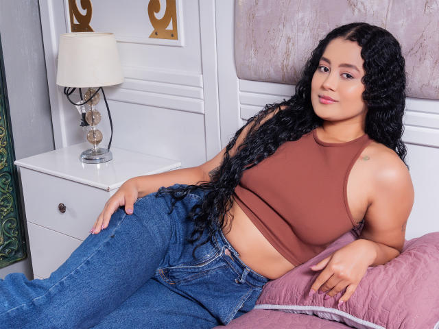 MaddiAndAndrew with curly black hair, brown crop top, and blue jeans, laying on pink pillow, smiling, showing small tits, and a cute butt, near white nightstand with lamp.