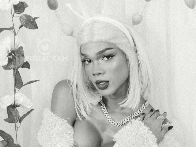 NaomiSteward with blonde bob, bunny ears, fluffy off-shoulder top, chain necklace, looking seductively at camera, gray-scale, revealing cleavage, slightly parted lips, watermark visible.
