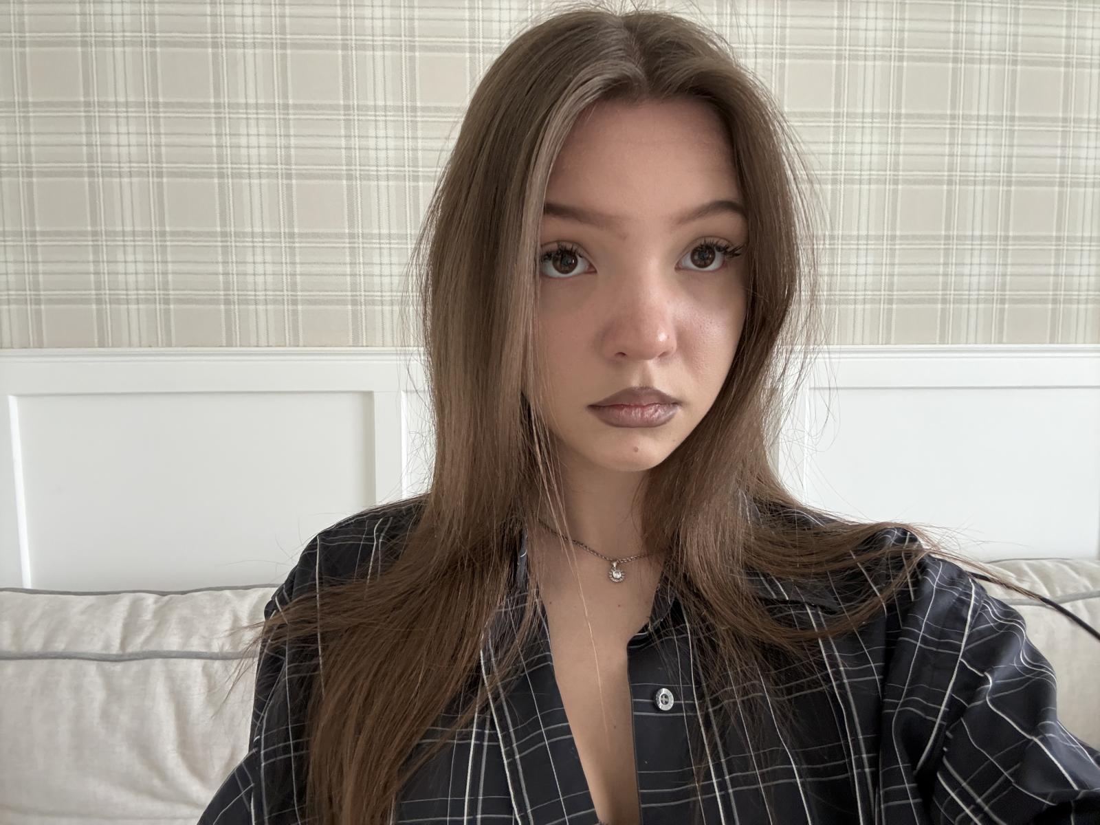 SondraKlebes has long, straight brown hair and a neutral expression. She's wearing a black plaid shirt with a small button near the collar, slightly open, revealing a hint of cleavage. She has a small silver necklace with a round pendant. Her brown eyes are slightly parted, and she has subtle eyeliner. The image has a realistic style, with natural lighting and a soft focus. She's centered in the frame, with a white headboard and a beige plaid pattern in the background.