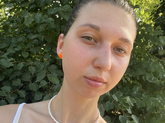 Sanella has a fair complexion, light brown hair pulled back, and green eyes. She wears small orange earbuds and a white necklace. Her face is slightly freckled, with natural, light pink lips. She has a subtle, calm expression. The photograph is close-up, focusing on her face and upper shoulders. The background consists of green, leafy foliage. The light is natural, highlighting her skin texture and the small, dark eyebrows. The image has a clear, realistic style.