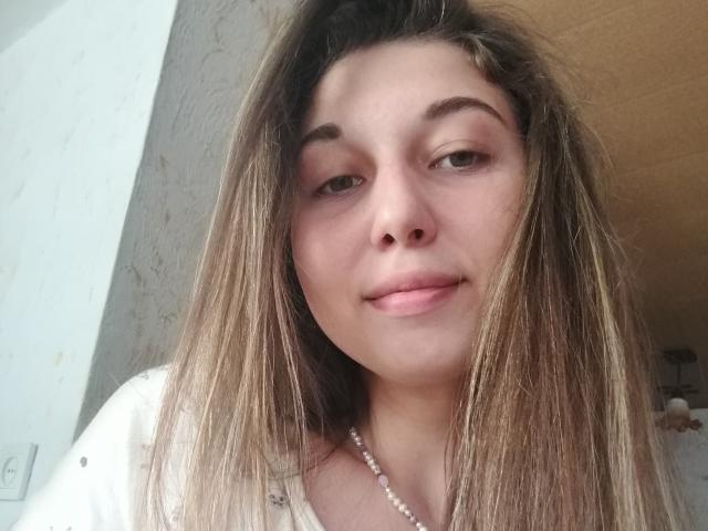Sanella has light brown, straight, medium-length hair, a fair skin tone, and a slight smile with pink lips. She wears a white shirt and a pearl necklace.