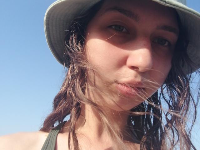 Close-up of Sanella with wavy brown hair, wearing a green tank top and a grey sun hat. She has light skin, green eyes, and slightly pouty lips. Her hair is blowing in the wind, and the sun is casting shadows on her face. The image is taken from a low angle, emphasizing her upper body and face. The clear blue sky forms the background. Sanella's expression is neutral, and her gaze is directed slightly to the right. Her eyebrows are thick and dark, contrasting with her lighter skin.