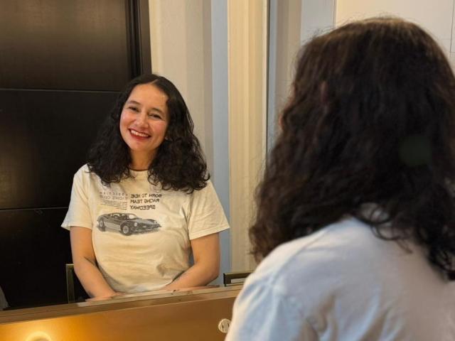 LilithCrowe with curly black hair, smiling at her reflection in a mirror, wearing a white t-shirt with a car graphic, standing in front of a black door.