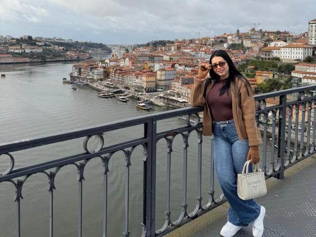 IsabellaJames stands on a bridge, wearing a brown jacket, dark turtleneck, blue high-waisted jeans, and white sneakers. She holds a white handbag and adjusts her sunglasses. Her long black hair flows down her back. She has a confident, relaxed posture. The bridge has ornate black iron railings. The background shows a bustling riverfront city with colorful, historic buildings and a cloudy sky. The river is calm, with boats docked along the waterfront. IsabellaJames's stylish, casual outfit contrasts with the city's architectural details.