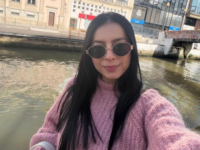 IsabellaJames stands by a river, wearing round, pink-framed sunglasses and a textured, pink sweater. She has long, straight black hair and a slight smile. The river's greenish water reflects sunlight. The background includes a mix of old and modern buildings, with a glass-fronted structure on the right. Her skin is light, and she has a relaxed, confident posture. The image is a clear, daytime selfie.
