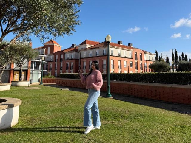 IsabellaJames stands in a grassy area, wearing a pink sweater, blue jeans, and white sneakers. She's holding a small object in her right hand. She's positioned slightly to the right, with a large, red-brick building and a tall lamp behind her. A tree is on the left, and a green, metal lamppost is in the middle. Clear blue sky and shadows are visible. Grass is well-maintained. She's facing slightly to the left.