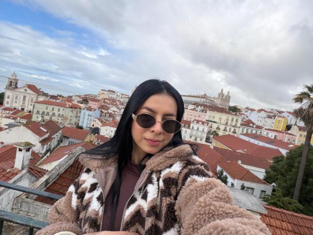 IsabellaJames with long black hair, brown eyes, wearing round sunglasses and a brown, white, and black fuzzy jacket, standing on a rooftop with a city skyline behind her.