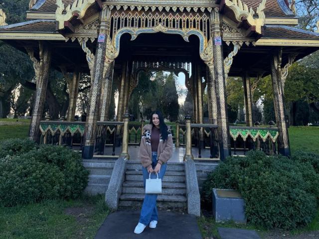 IsabellaJames stands in front of an ornate, gold-trimmed wooden pavilion. She has long dark hair, a brown jacket, blue jeans, and white shoes. She holds a white bag and a coffee cup with a smile. The pavilion has intricate patterns and green trim. Bushes frame the steps where she stands. The image is a clear, detailed photograph with natural light.