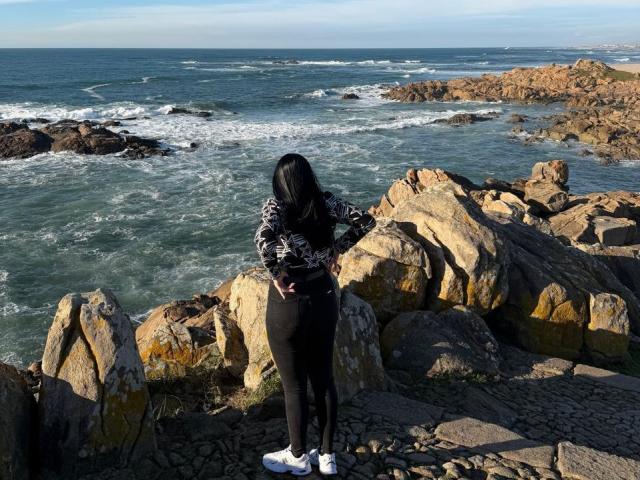 IsabellaJames stands on rocky coastline, facing ocean. She has long black hair, wearing black patterned sweater, black pants, white sneakers. Rocks in foreground are large, jagged, sunlit. Ocean waves crash against rocks in midground. Sky is clear with light blue gradient. IsabellaJames's hands rest on her hips. Composition focuses on contrast between her dark clothing and bright rocks. Natural light highlights textures. IsabellaJames is centered, facing right.