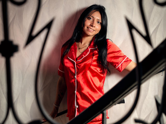 VioletXBloom stands against a textured wall, wearing a shiny red satin dress with white trim. She has long black hair and a slight smile, looking directly at the camera. The dress contrasts with the white wall, and a black, geometric-patterned frame is in the foreground, slightly out of focus. Soft lighting highlights her face and dress, creating subtle shadows. Her right arm rests on a black railing.