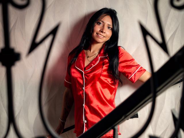VioletXBloom stands in a red satin shirt with white trim, leaning against a textured white wall. Her long black hair is loose, and she smiles slightly. She wears a necklace and has tattoos on her left arm. The black metal railing in the foreground creates a shadow over her face. The red shirt contrasts with the white wall and her dark hair. The image uses soft lighting to highlight her face and the satin texture.