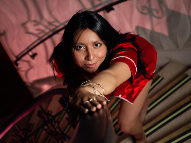 VioletXBloom is a young Asian woman with long black hair, wearing a red satin dress with white trim. She's leaning forward over a dark wooden railing, looking directly at the camera with a slight smile. She has brown eyes and a light skin tone. Her right arm is extended, with her hand gripping the railing, and she wears a silver bracelet and a ring on her right hand. The lighting casts a pinkish shadow on the wall behind her. The stairs are striped in green and beige. The composition is tight, focusing on her upper body and face.