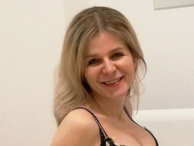 VeraBuck77-hot with blonde hair, smiling, wearing a black strappy top, showing her small tits and cleavage, in a selfie with soft lighting.