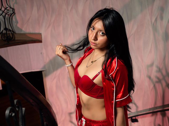 VioletXBloom has long black hair, brown eyes, and a medium skin tone. She wears a red satin bra and a matching red satin jacket with white trim, open to reveal her bra. She holds a strand of her hair with her right hand, looking slightly to the side. She has a necklace with a small pendant and a white bracelet on her right wrist. The background has a textured pink wall and a wooden staircase with a metal railing. The lighting is soft, with shadows on the wall.