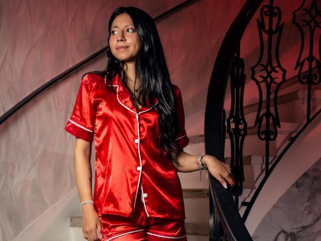 VioletXBloom stands on a staircase, wearing a shiny red satin pajama set with white trim. Her long black hair is straight, and she looks to the side with a subtle smile. She has a light brown skin tone and a slim build. She rests her right hand on the black, ornate railing. The red satin contrasts with the white staircase and the slightly textured wall behind her. She wears a silver bracelet on her right wrist. The lighting has a red tint.