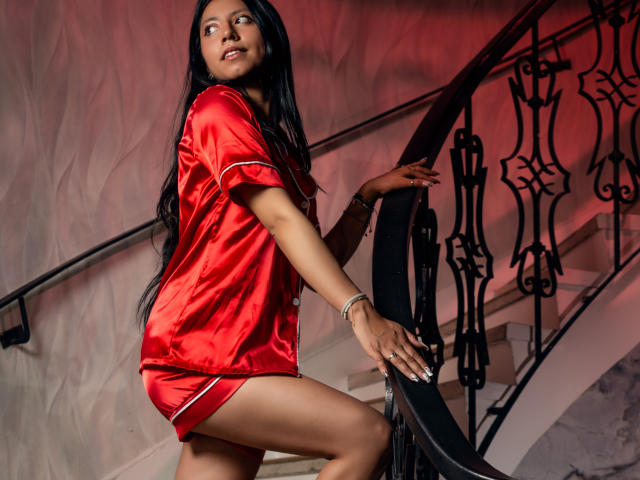 VioletXBloom stands on a staircase, wearing a shiny red satin dress with white trim. Her long black hair is straight, and she looks up with a slight smile. Her right hand grips the black ornate railing, while her left rests on it. She has dark skin, and her nails are painted white. Red lighting casts a warm glow on the white wall behind her. The staircase has a curved design with black metal railings. Her dress is short, revealing her thighs. She wears a silver bracelet on her left wrist.