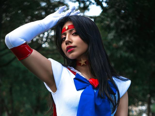 VioletXBloom is a woman with long black hair, wearing a red headband with a gold star, white gloves, and a white and blue costume with a red bow. She has red lipstick and a star-shaped choker. Her right arm is raised, hand touching her head. The costume has a red chest bow and blue necktie. The image has a green, blurred forest background. The photo is bright and focused on VioletXBloom.