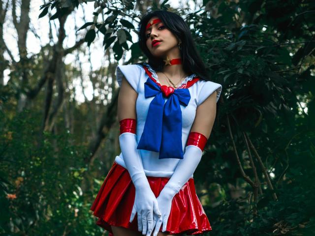VioletXBloom stands in a forest, dressed in a Sailor Moon cosplay outfit. She wears a white, sleeveless top with a large blue bow at the chest, red skirt with a shiny texture, white gloves, and red arm bands. She has long black hair, red eye mask, and red lipstick. Her hands are clasped in front of her. The background is dense with green foliage. The image has a vivid, high-contrast style with a focus on VioletXBloom's colorful outfit against the natural greenery.