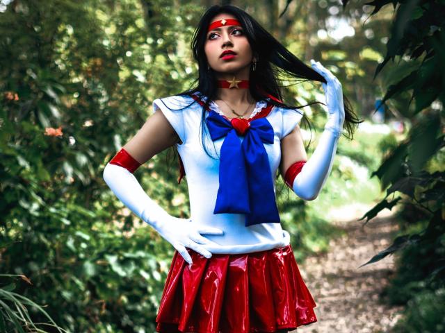 VioletXBloom stands in a forest, wearing a Sailor Moon costume. She has long black hair, red eyes, and red lipstick. She wears a red headband, white gloves, a blue bow, and a red skirt with a shiny texture. Her right hand holds her hair. The costume features a white top with red and blue accents. The forest is green and blurry in the background. The image is colorful and detailed.