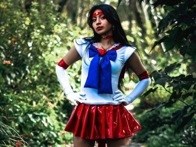 VioletXBloom stands in a forest, wearing a Sailor Moon cosplay outfit. She has long black hair, red lipstick, and a red headband. Her outfit includes a white top with a blue bow, red skirt, white gloves, and red arm cuffs. Her hands are on her hips, and she looks forward with a serious expression. The forest background is blurred, highlighting her in focus. The colors are vivid, with bright reds, blues, and whites contrasting with the green foliage. The image is sharply detailed, emphasizing her cosplay attire.