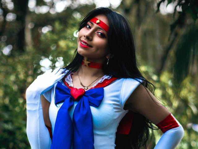 VioletXBloom in a Sailor Moon cosplay outfit. She has long black hair, a red headband, red lipstick, and a red choker. She wears a blue and white costume with a large blue bow, a red heart-shaped chest piece, and white gloves. Her right hand is raised, touching her hair. She has a slight smile. The costume is shiny with red and blue fabric. Her skin is light. The image is focused on VioletXBloom with a blurred forest background.