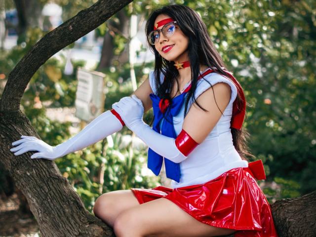VioletXBloom in a detailed Sailor Moon cosplay, sitting on a curved tree branch. She wears a white sleeveless top with a blue bow and red skirt, red and blue gloves, and a red headband with a yellow star. Her long black hair is loose, and she has red lipstick. The bright red and blue colors contrast with the green foliage in the background. The image is a vivid, realistic photograph.
